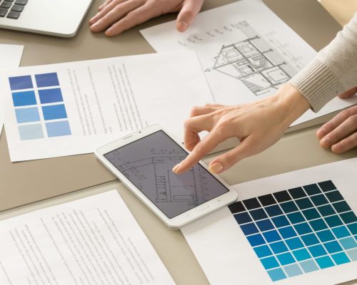 Close up image of house plan drawings, pantone swatches on desk, businesswoman tapping digital tablet screen with apartment floor scheme presenting construction project to a partner. Teamwork concept
