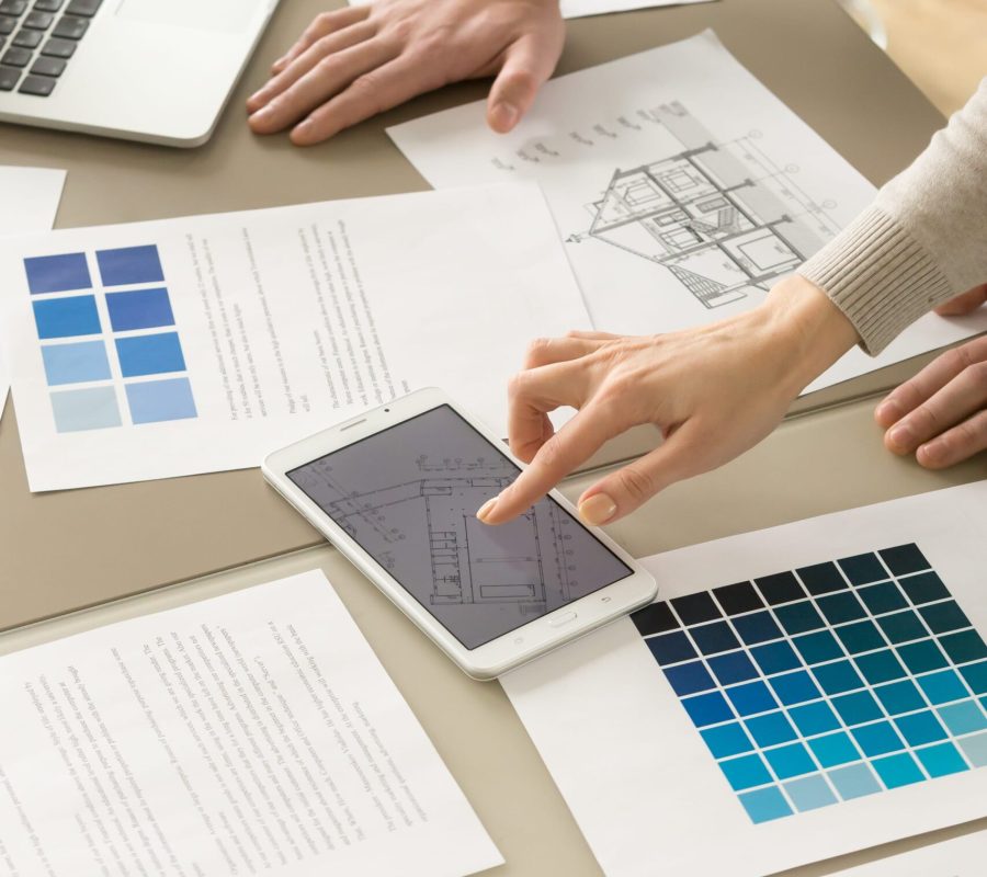 Close up image of house plan drawings, pantone swatches on desk, businesswoman tapping digital tablet screen with apartment floor scheme presenting construction project to a partner. Teamwork concept